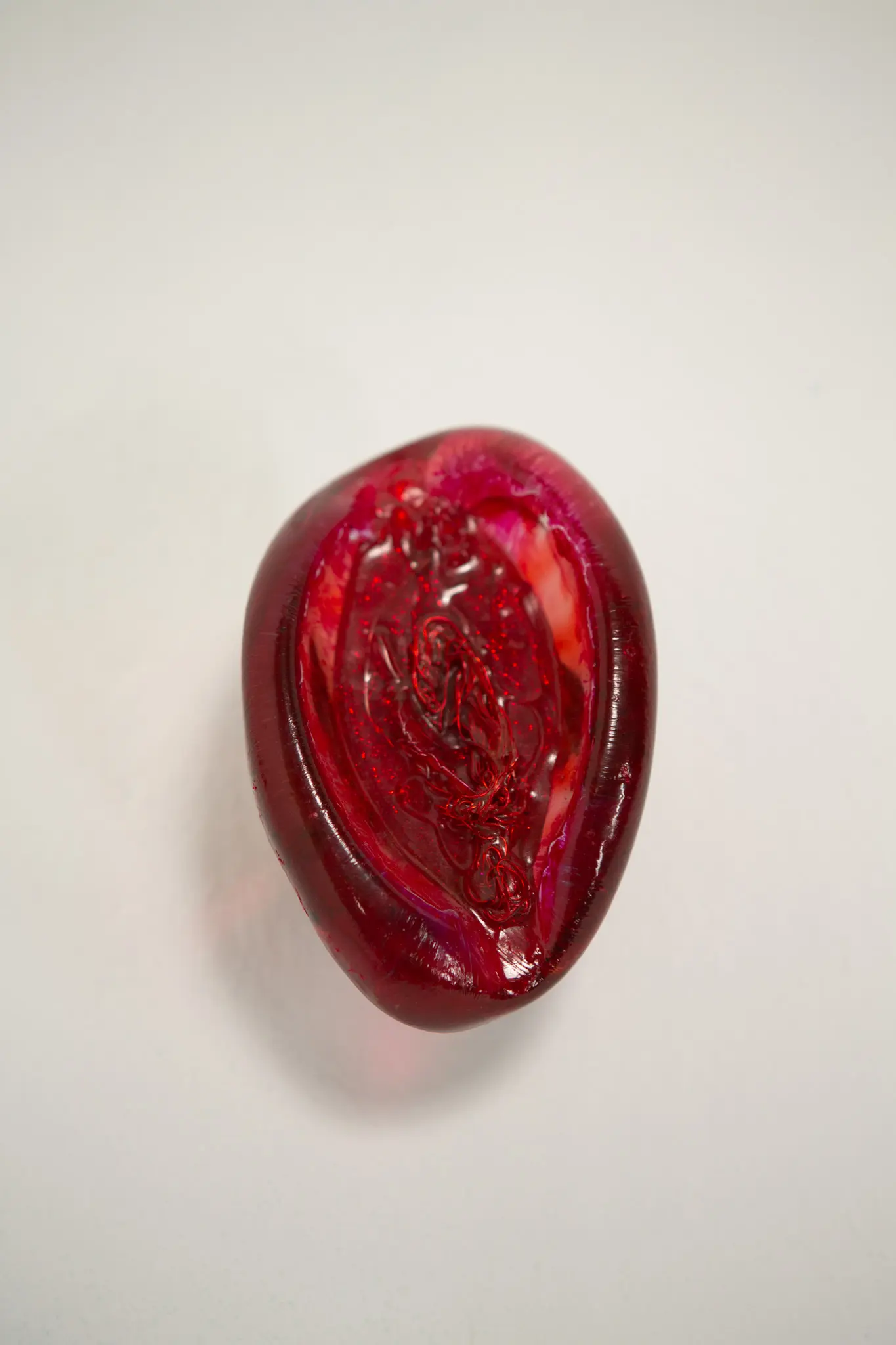 Forme verticale en resine cramoisie vue de face, creux interieur a paillettes enserrant un fil de fer rouge, posee sur fond blanc casse.