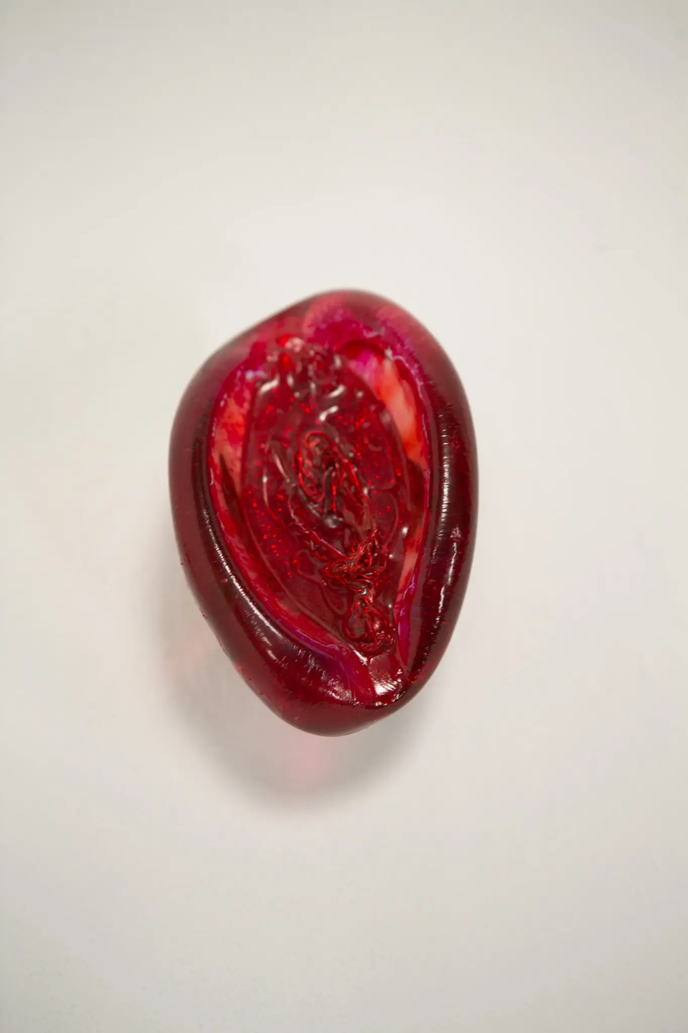 Objet en resine rouge en amande, surface luisante et cavite paillettee traversee d'un fil rouge, vue rapprochee sur fond neutre.