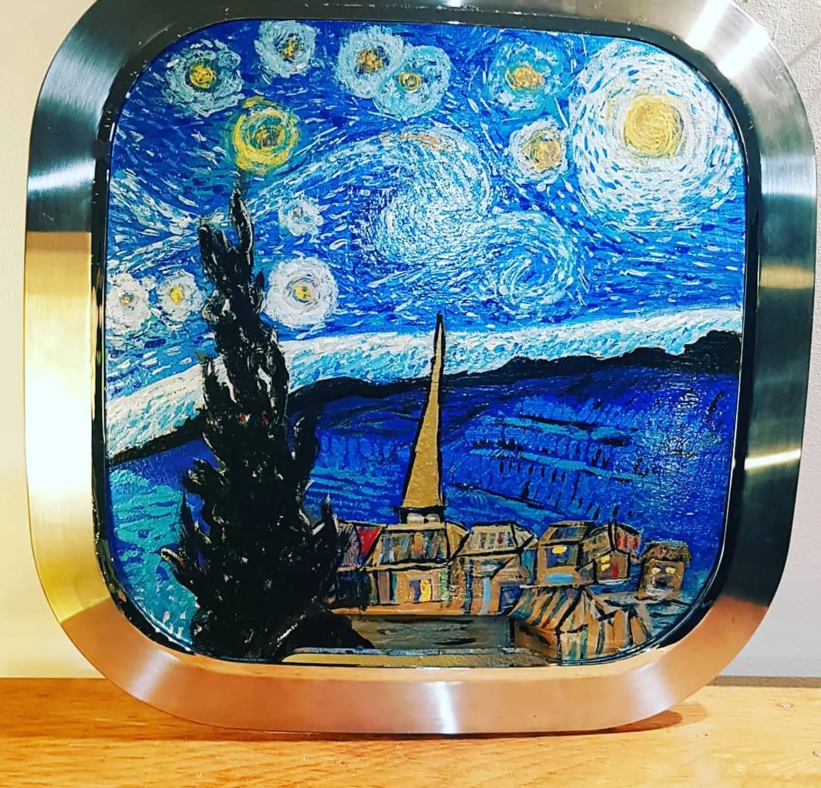 Peinture d'un ciel nocturne bleu tourbillonnant semé d'étoiles cernées de jaune, un cyprès noir devant un village au clocher ocre, sur une vitre bombée cerclée d'acier brossé aux angles arrondis, dressée sur une planche de bois clair devant un mur pâle