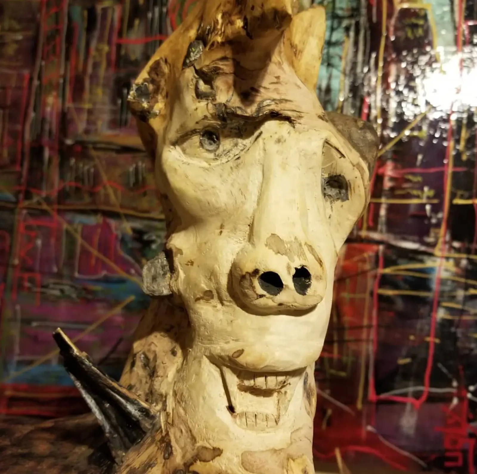 Gros plan serré sur une tête de bois pâle et mat, dont le sommet sort du cadre, museau percé de deux narines noires au-dessus d'une rangée de dents entaillée, éclats de pierre pris dans la matière, devant une toile peinte rouge et violet