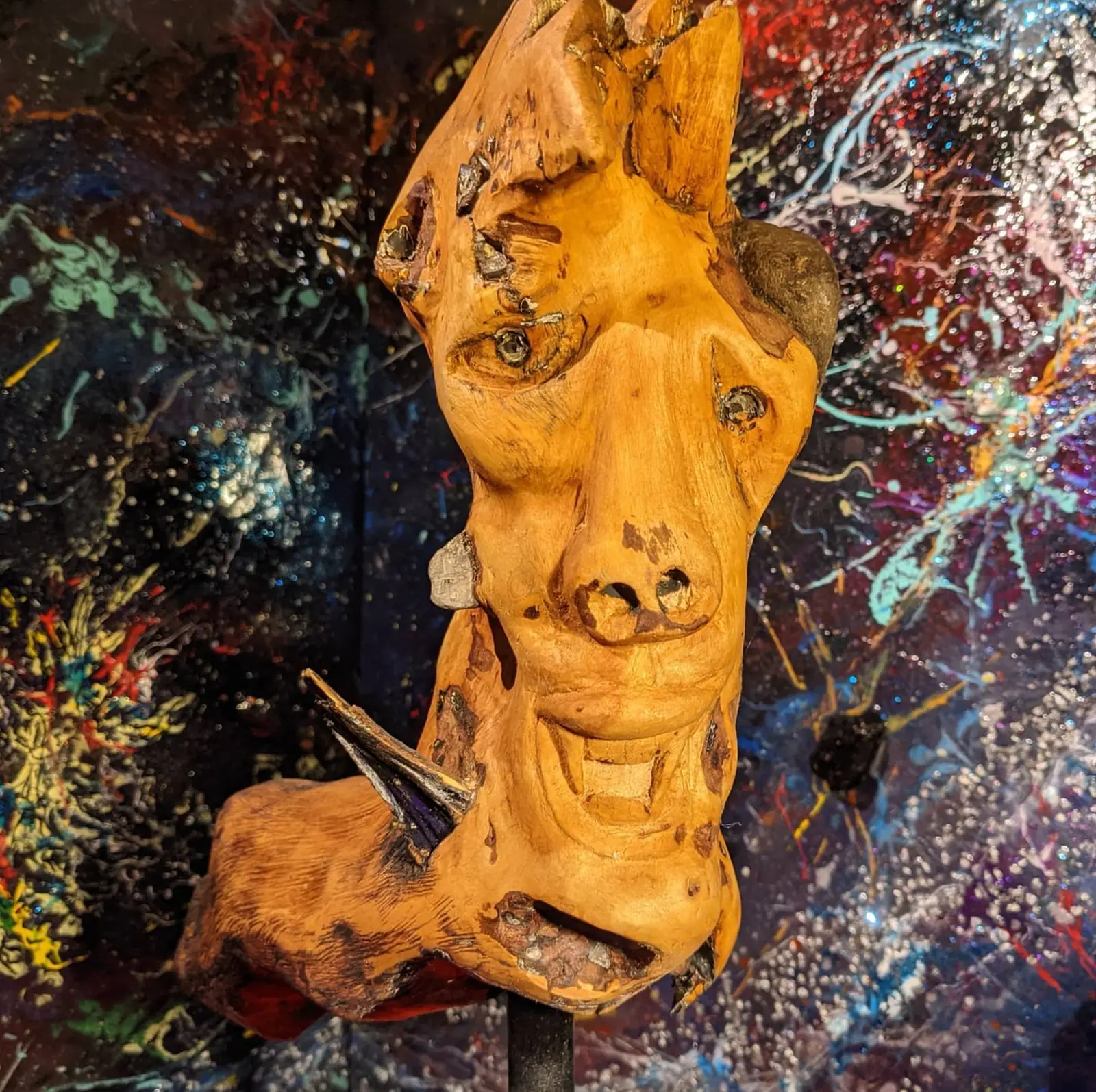 Vue de face de la tête en bois ambré et luisant, une pierre grise coincée sur son côté droit, le bois se prolongeant vers la gauche en une masse sombre, plantée sur une tige d'acier noire devant une toile éclaboussée de bleu et d'argent