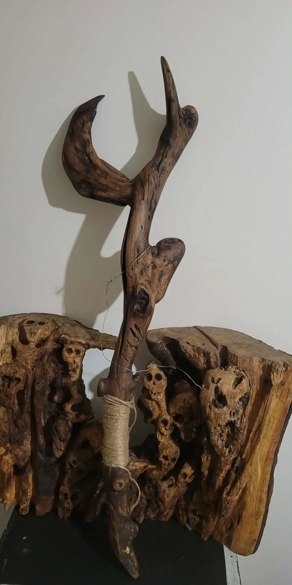 The Flipping Duck : branche de bois trouvé dressée, bec pointé vers le haut, corde de jute enroulée à la base, posée devant un panneau de bois sculpté de crânes.