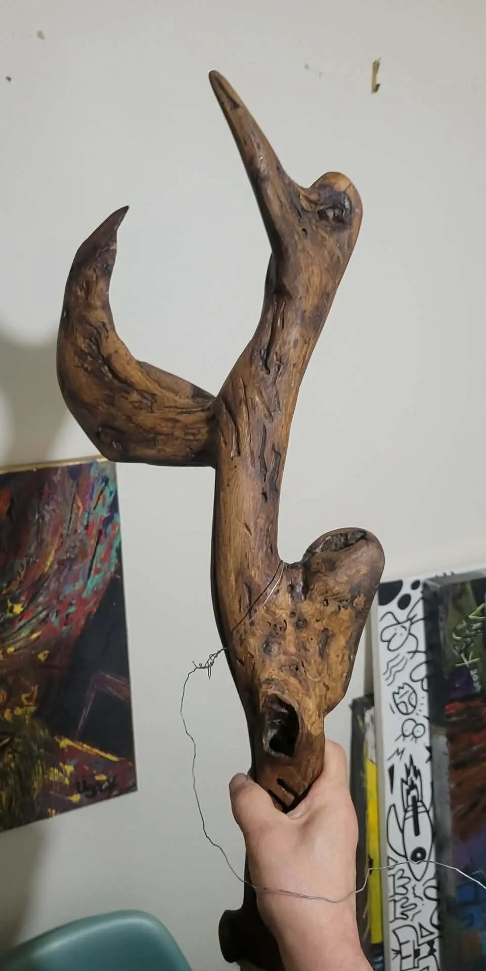 The Flipping Duck tenue à la main : la branche de bois trouvé vue de près, bec dressé, aile courbe et fil de métal, devant deux toiles.