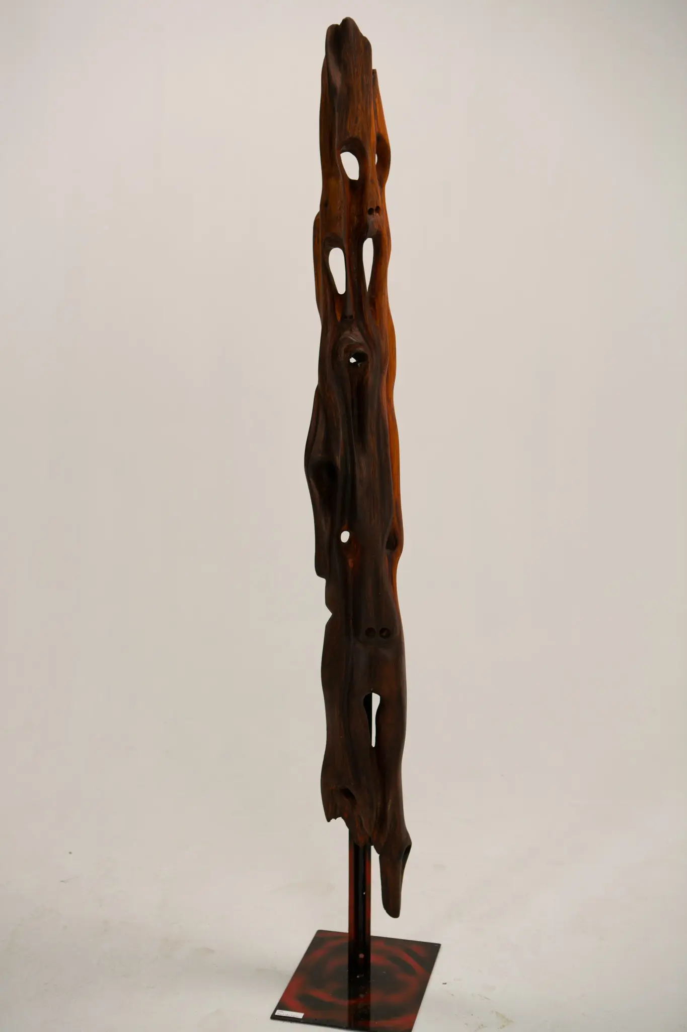 Sculpture verticale en bois trouvé à finition rouge-brun, percée de cavités sur toute sa hauteur, dressée sur une tige métallique et un socle carré au motif rouge sombre.